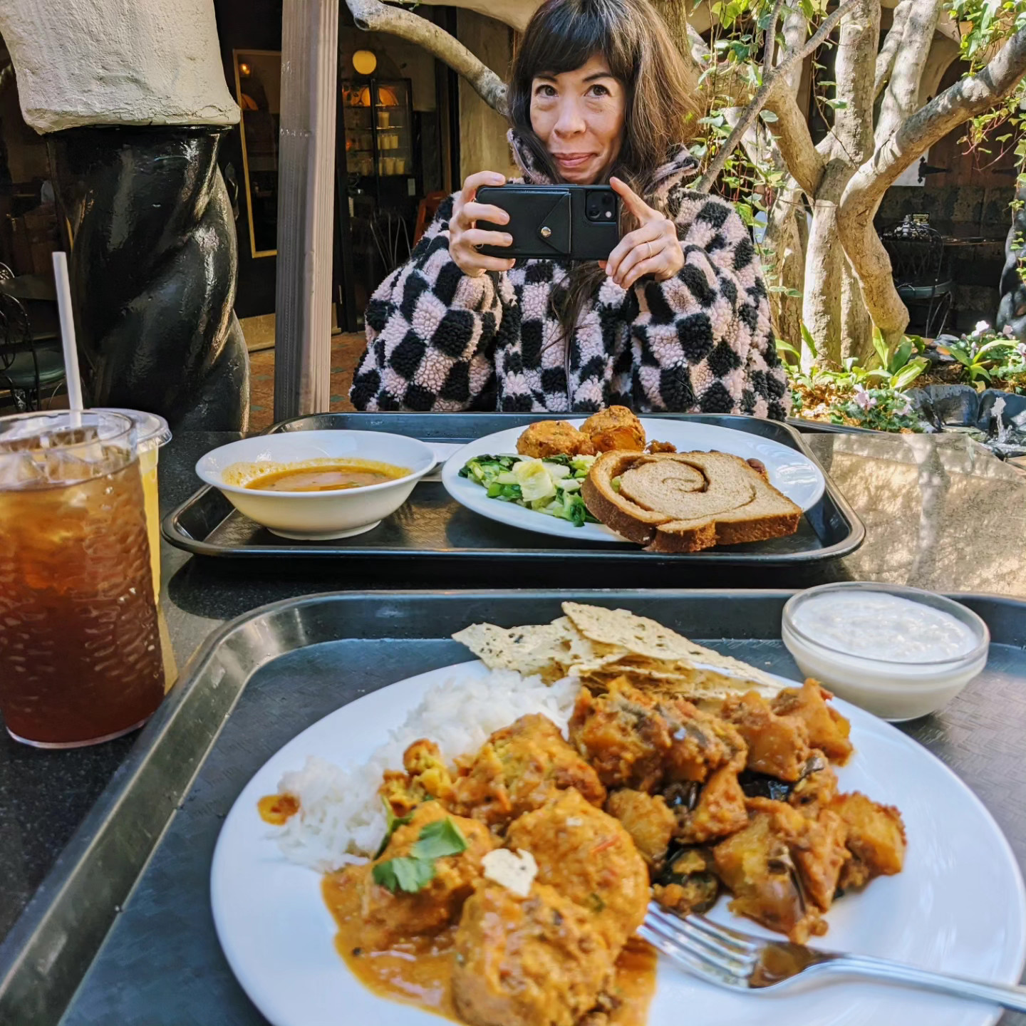Photo of a lunch. Two delicious looking plates of veggie Indian food. A woman sitting across the table talking a picture with her phone