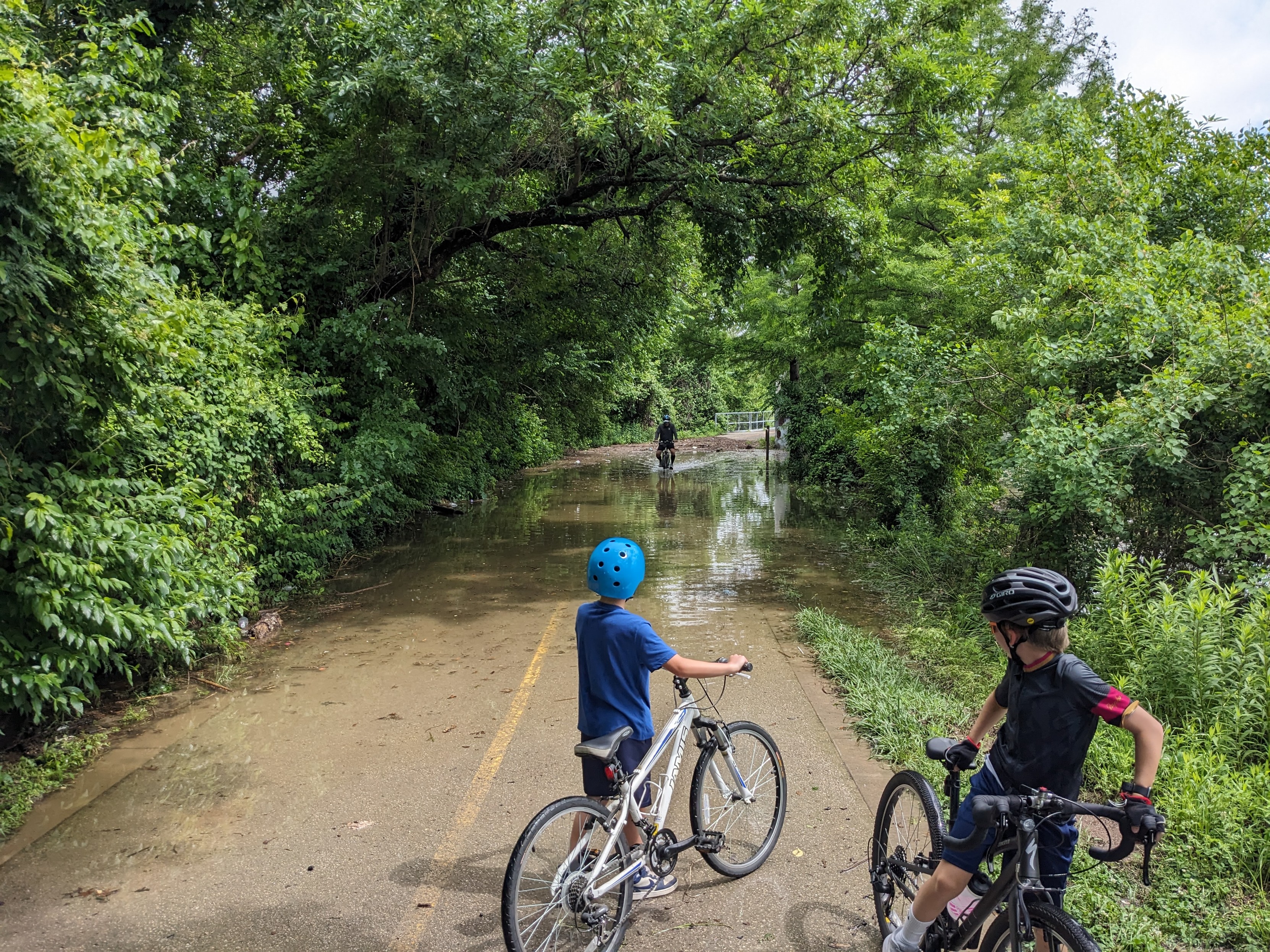 Two boys on bicycles looking at a flooded portion of bike trail. There is a cyclist riding through the flooded portion towards us.