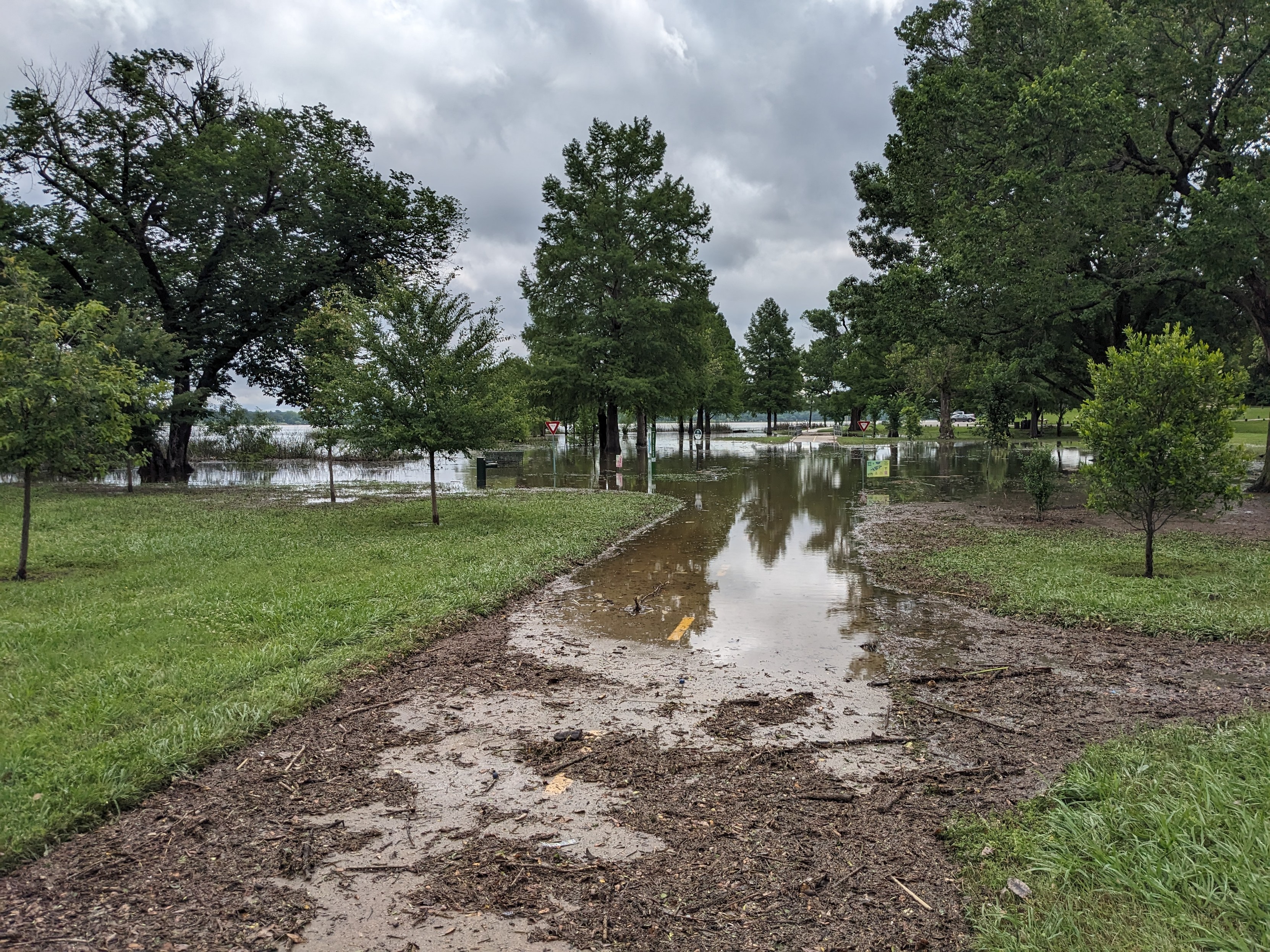 A very flooded portion of bike trail near a lake. There are piles of washed up dirt in the foreground where the water has receded a little bit.