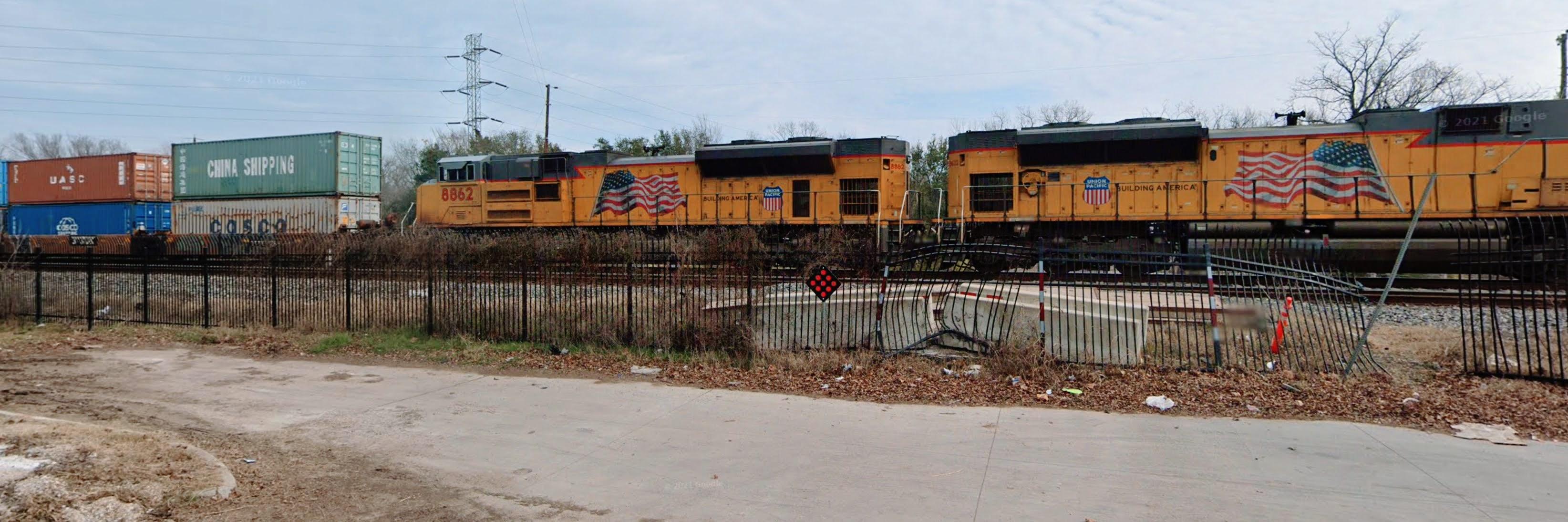 a railway scene with a Union Pacific freight train passing by. The locomotives are yellow and prominently feature the American flag and the words "Building America." Behind the engines, there are stacked intermodal containers labeled with brands like "CHINA SHIPPING" and "COSCO." The scene includes a black metal fence, part of which is bent and damaged, and a ground area littered with debris.