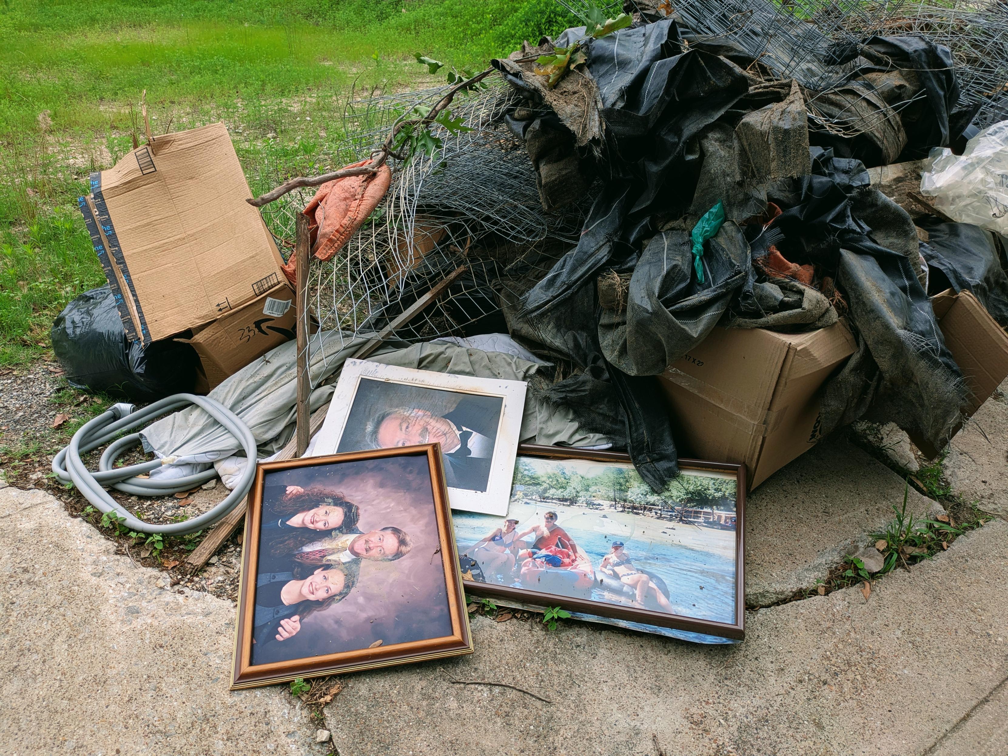 a pile of discarded household items on a curb, likely intended for trash or bulk pickup. The pile includes:

Torn and bundled black plastic sheeting and rolls of fencing or netting

Several cardboard boxes, some flattened

A black garbage bag

Coiled gray plastic tubing or hose

Framed photographs, including:

A posed family portrait of four people

A formal portrait of a man in a tuxedo

A casual group photo of people enjoying a boat or beach outing
