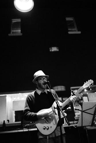 b/w photo of the author in 2008, playing an electric guitar and singing into a microphone at a nondescript location