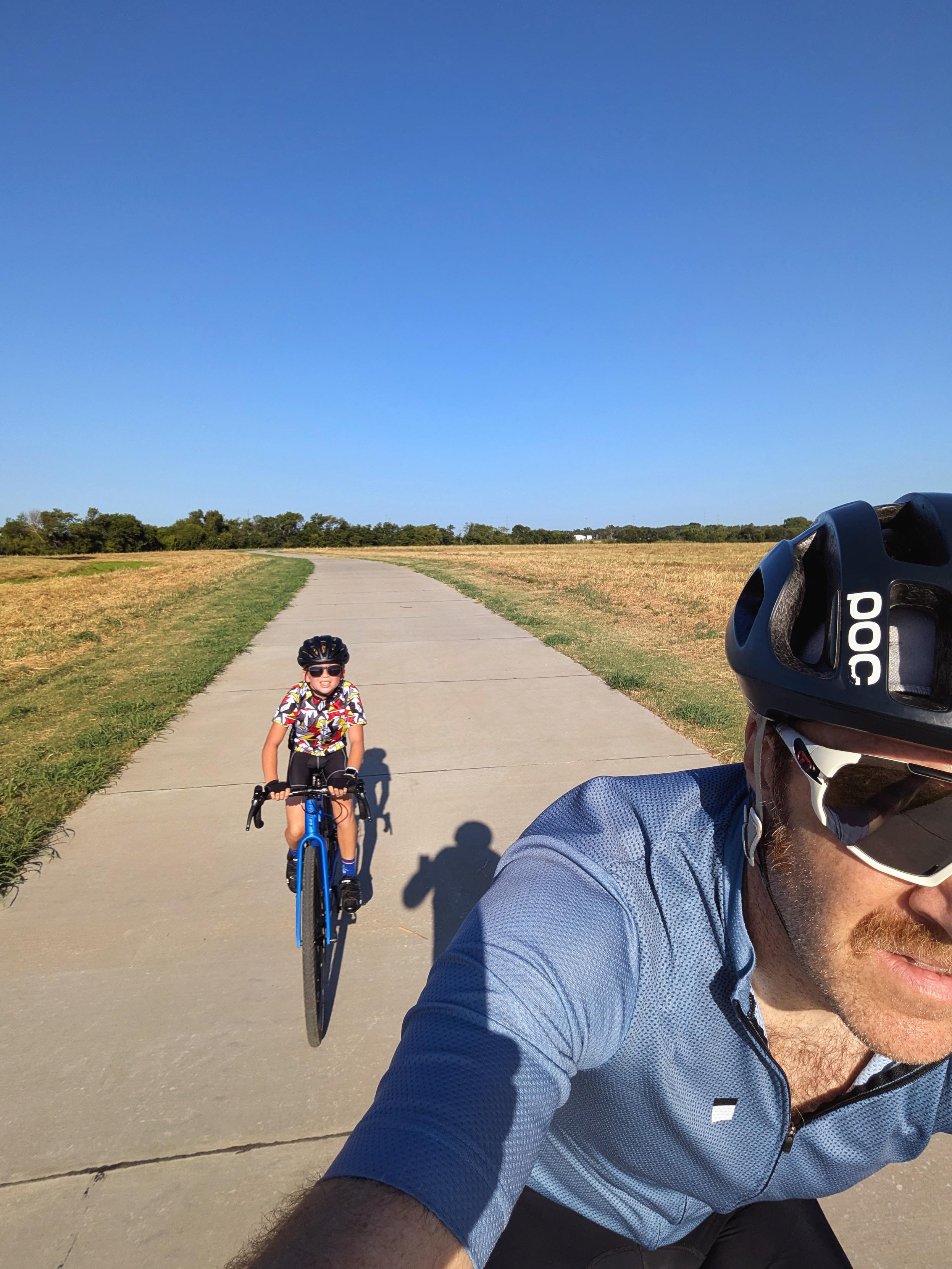 Bike selfie: half of a man seemingly on a bicycle in the foreground, boy behind him on a blue bicycle. The trail disappears behind them into the horizon, fields of brown grass on either side of the trail.