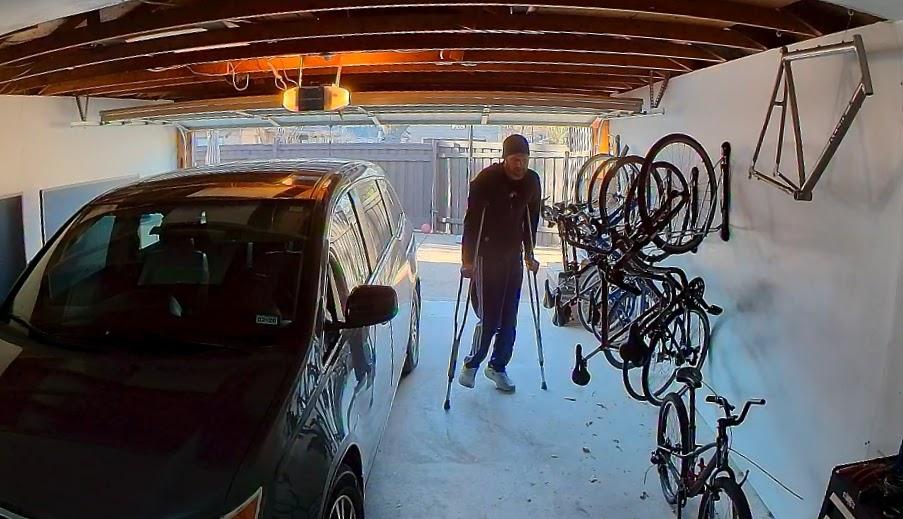 A person using crutches walks through a residential garage. A minivan is parked on the left, while several bicycles hang on wall-mounted racks to the right. The garage door is open, letting in daylight and revealing a fenced yard outside.