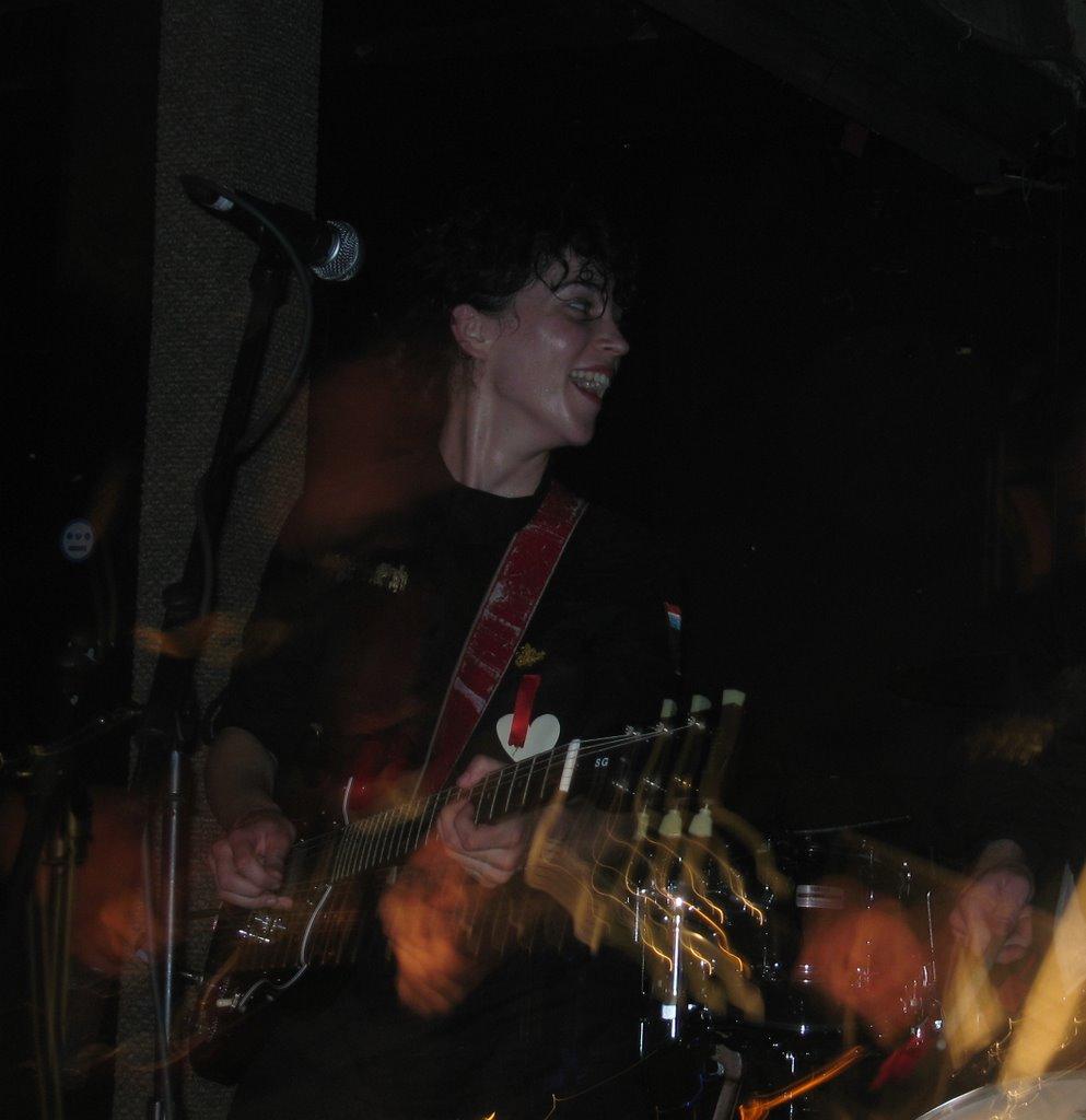 A dark, grainy concert photo of a woman (Annie Clark, aka St Vincent) playing guitar. She is wearing all black (the Polyphonic uniform from the Fragile Army era, although this show was from before the record release) and looking behind her, with a big smile on her face.