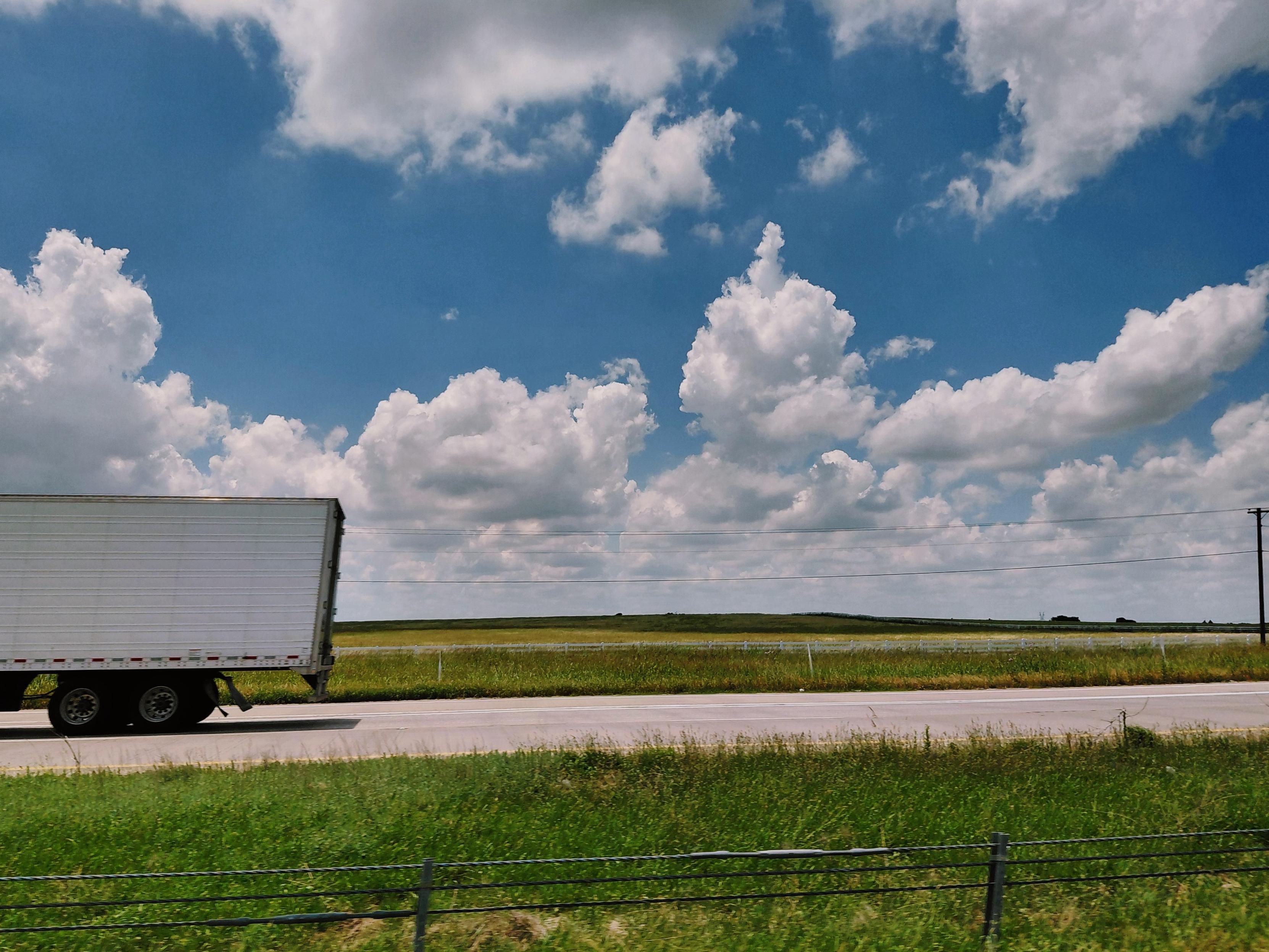 Photo from car window across opposite highway lane, green fields to horizon, big blue sky filled with fluffy white clouds. The back of a tractor-trailer on the left.