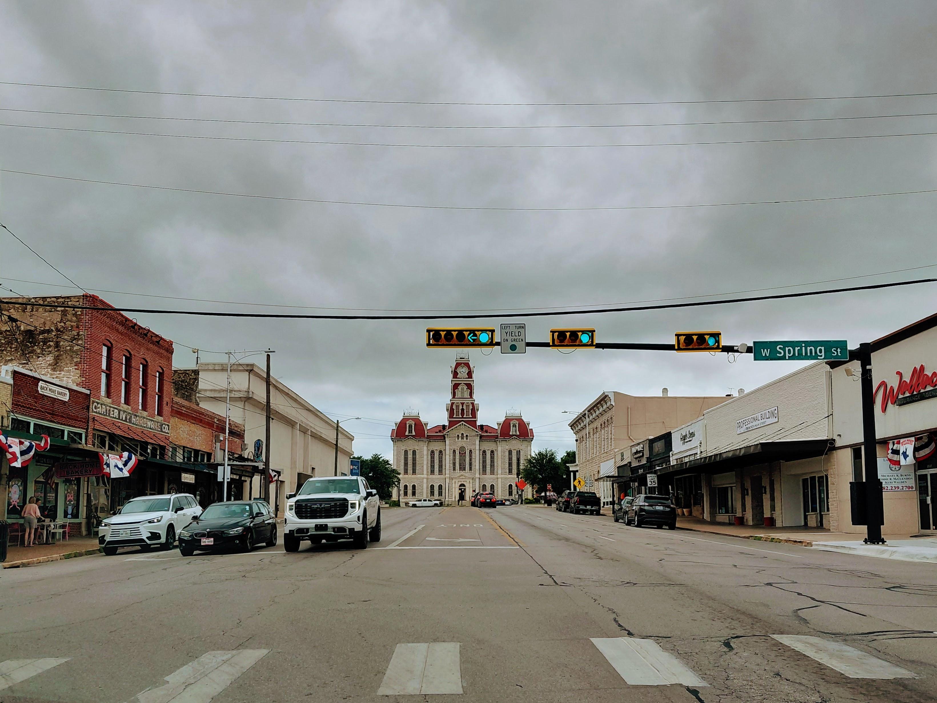 A two-lane small-town main street stretches toward a historic courthouse with a red-roofed clock tower centered in the distance. Storm clouds fill the sky overhead. Traffic lights hang across the intersection, and a green street sign reads “W Spring St.” Brick storefronts and parked vehicles line both sides of the street, with faded patriotic bunting decorating some of the buildings. The scene is viewed from the middle of the road, emphasizing the courthouse as the focal point.