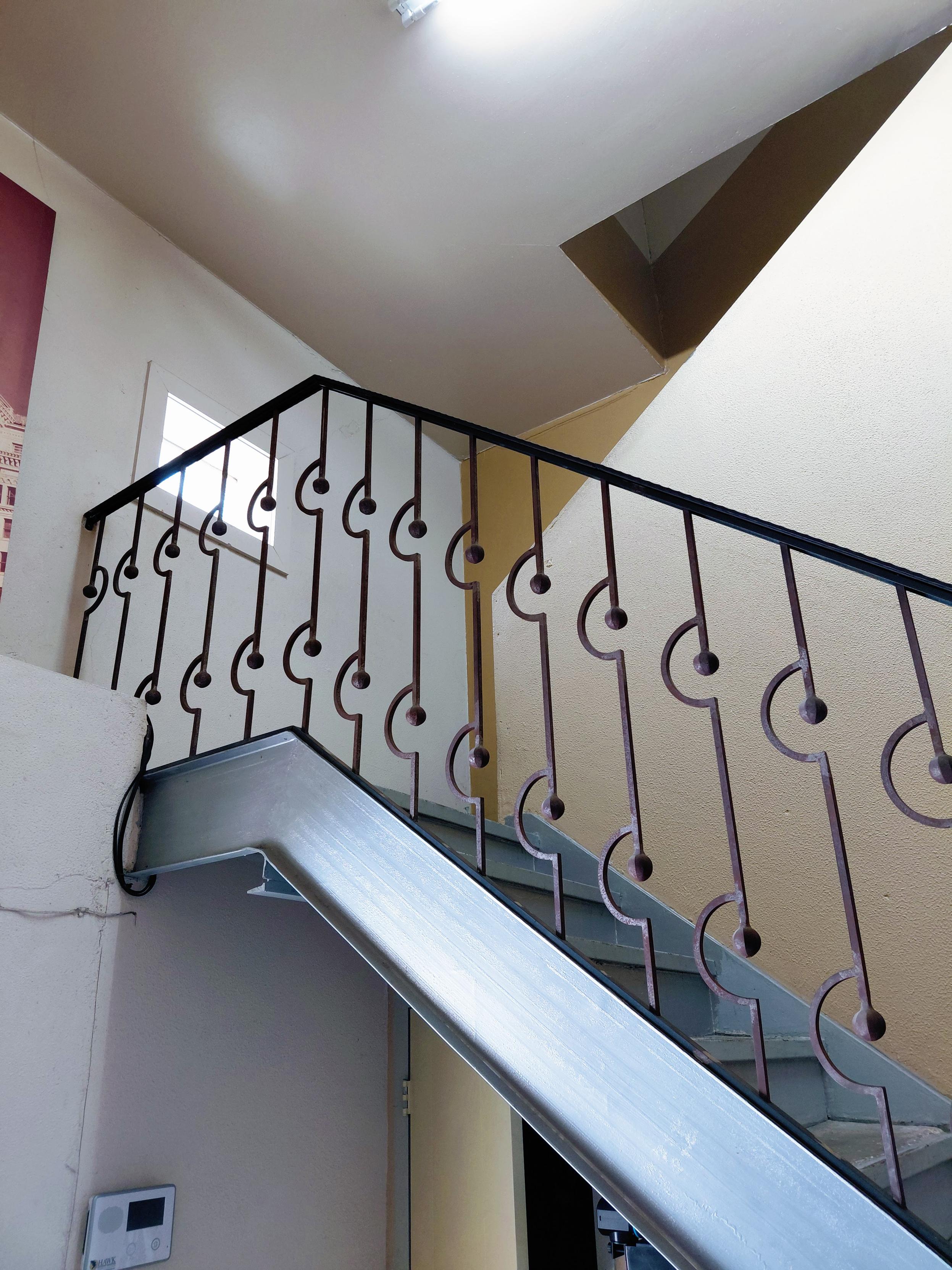 Interior stairwell with gray metal stairs and a decorative metal railing featuring repeating curved and circular designs. White and beige walls, a small window, and an angled ceiling frame the space, with daylight and a ceiling light illuminating the scene.